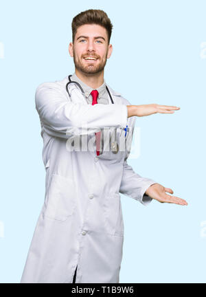 Young handsome doctor man wearing medical coat gesturing with hands showing big and large size sign, measure symbol. Smiling looking at the camera. Me Stock Photo