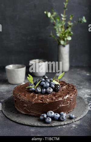 Tasty dark chocolate cake with blueberries and green leafs, two cup of tea, vase with herbs on gray background. Concept of birthday chocolate cake. De Stock Photo
