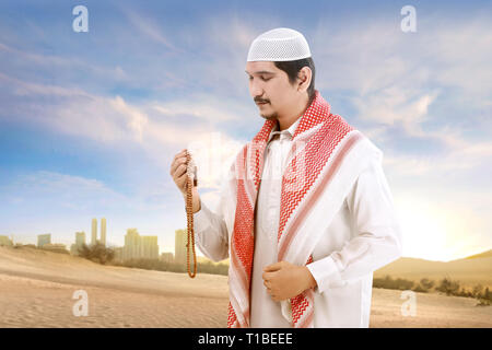 Young asian muslim man with cap and turban standing and praying with prayer beads on the sand with blue sky background Stock Photo