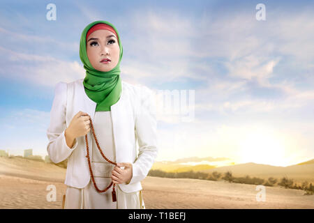 Young asian muslim woman in veil standing and praying with prayer beads on the sand with blue sky background Stock Photo