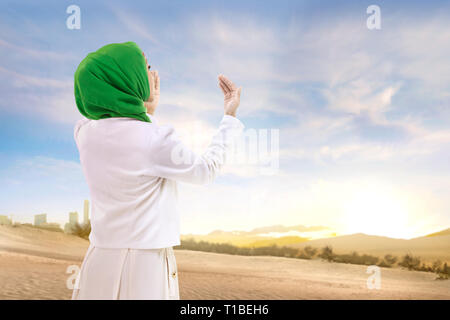 Beautiful asian muslim woman in veil standing and praying with raised arms on the sand with blue sky background Stock Photo