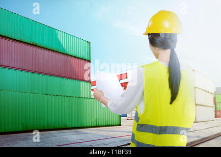 Rear view of asian worker woman with yellow hard hat checking the shipping schedule on the industrial harbor with blue sky background Stock Photo