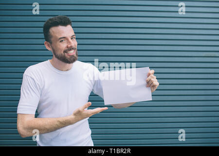 Nice and positive man iss standing and smiling. He is posing and looking at camera. Guy is holding piece of paper and poiting on it with hand Stock Photo