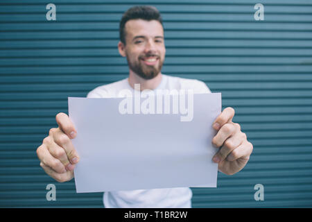 Nice and good-looking man is looking on camera. He is holding blank piece of paper with both hands. He is showing it to camera. Isolated on striped Stock Photo