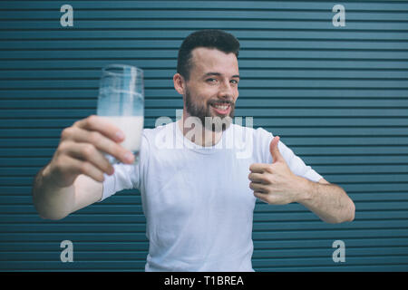 Nice and positive man is holding glass of mils and showing big thumb up. He likes it. Guy is looking on camera and smiling. Isolated on striped and Stock Photo