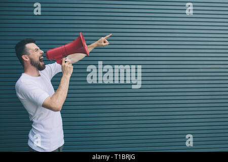 A picture of man standing and pointing forward. He is talking and screaming into megaphone. Isolated on striped and blue background. Stock Photo