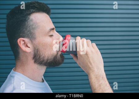 Guy is drinking coffee from plastic cup. He is keeping eyes closed. Isolated on striped and blue background. Stock Photo