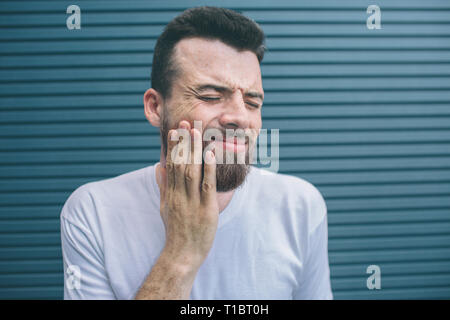 young man is standing and holding hand on cheek. He is keeping eyes closed. Guy feels pain. Isolated on striped and blue background. Stock Photo