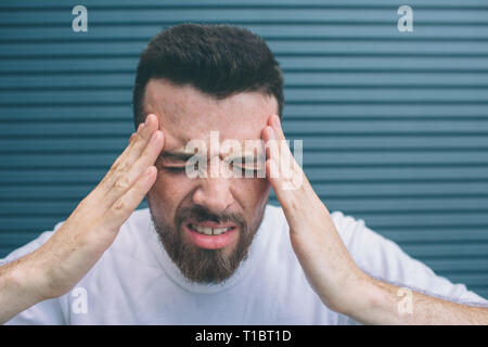 Young man is holding hands on forehead. He has headache. Guy feels terrible. He is suffering. Isolated on striped nad blue background. Stock Photo