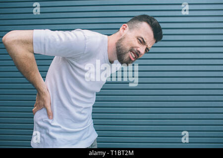 Unhappy guy is suffering from back pain. He is holding hands on spine and keeping eyes closed. Isolated on striped and blue background. Stock Photo