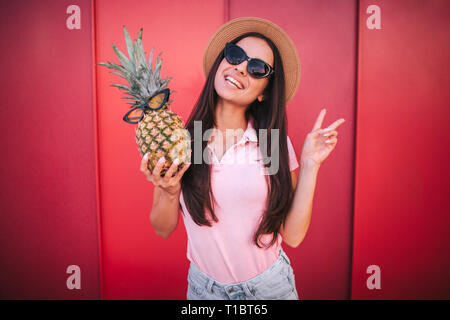 Beautiful woman stands and poses on camera. She shows the piece symbol with her fingers. Woman has pineapple in right hand. Both she and the fruit Stock Photo