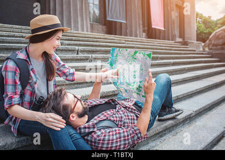 Nice picture of young tourists on stairs. She sits there and points on map. He holds map and lies on woman's knees. Stock Photo