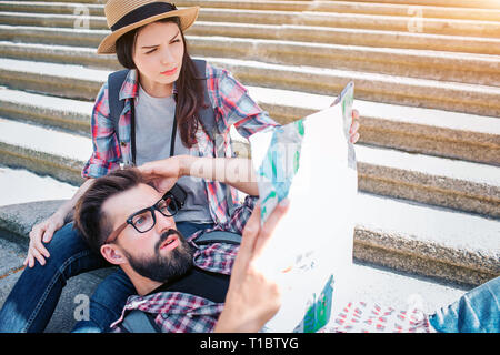 Picture of serious tourists on steps. He is lying on girlfriend's knees and hold map in hanad. She sits on steps. They are confused and concentrated. Stock Photo