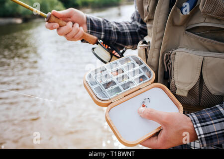 Close up of wooden box with diffeent kind of artificial silicone fishing flies and baits in it. Man holds it in one hand and has rod in the other one Stock Photo