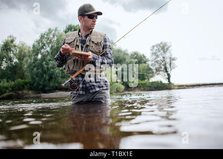 A picture of man holding box with flies and plastic artificial baits. He is holding one of them in hand. Also guy has a fly rod. He is holding it with Stock Photo