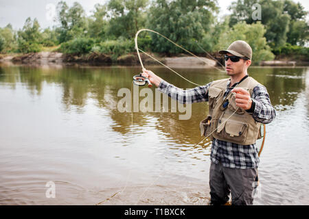 Serious fisherman stands on shallow an waving with fly-fishing. Also he has a spoon in other hand. Guy is looking to the left. He is concentrated. Stock Photo
