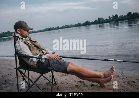 MAN SITTING IN CHAIR HOLDING FISHING ROD WITH TACKLE BOX AND NET