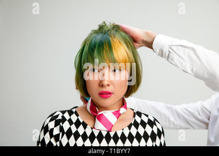 Picture showing adult woman at the hair salon. Studio shot of graceful young girl with stylish short haircut and colorful hair on gray background. Hands of hairdresser close up. Stock Photo