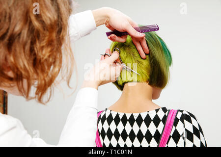 Picture showing adult woman at the hair salon. Studio shot of graceful young girl with stylish short haircut and colorful hair on gray background and hands of hairdresser. Stock Photo