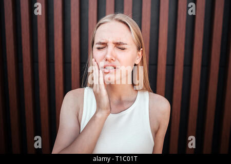 Beautiful blonde teen is standing and holding hand on cheeck. She has a tooth pain. Girl is upset. She is suffering. Isolated on striped background. Stock Photo