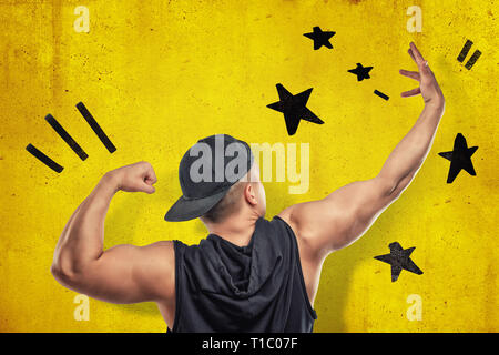 Strong muscular young man showing biceps with black stars drawn on yellow wall background Stock Photo