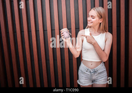 Positive blonde girl is looking on a jar of dessert she holds in hand. She is pointing on it. Young woman looks happy. Isoated on striped background. Stock Photo