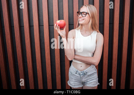 Healthy girl is holding red apple in hand and looking on it. She is smiling. Isolated on striped background. Stock Photo