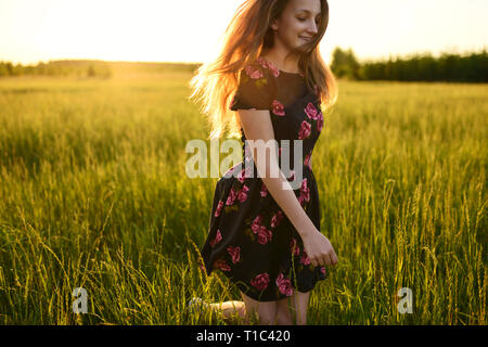 Young girl in flower dress, is running or jumping from happiness on a green meadow. She is smiling and her long hair are flowing on wind, all lit by w Stock Photo