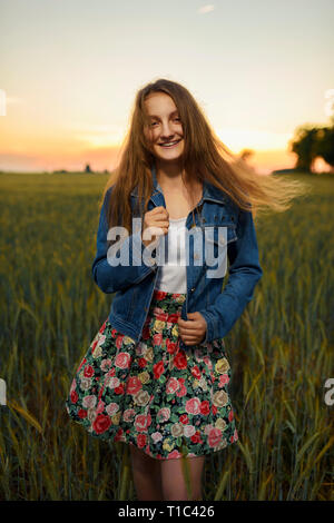 Smiling pretty girl in flower print skirt and jeans jacket, standing on a green cereals field crops, with warm sunlight of sunset. Countryside scenery Stock Photo