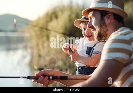 A mature father with a small toddler son outdoors fishing by a lake. Stock Photo