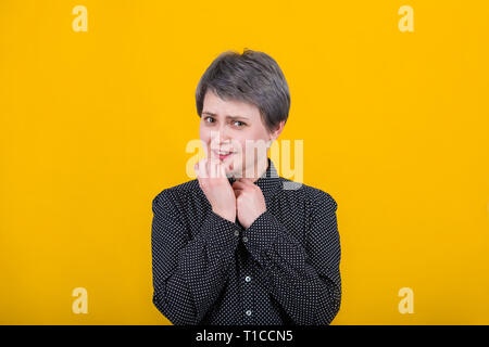 Scared and skeptical woman, keeps hands under chin, sees something terrible isolated over yellow wall. Human face expressions and emotions concept. Af Stock Photo