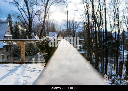 A snow-covered, wooden balustrade on the balcony with selective focus. In the background, blurred buildings and trees. Stock Photo