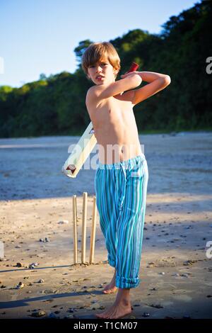 Handsome young 8-10 year old boy having fun playing cricket with bat on the beach Stock Photo