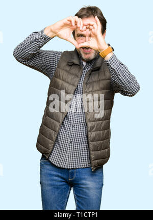 Middle age handsome man wearing winter vest Doing heart shape with hand and fingers smiling looking through sign Stock Photo