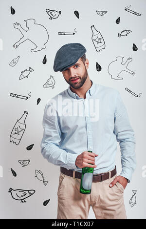 Bad boy. Bearded brutal man in hat holding a beer and looking ar camera while standing against grey background with hand drawn doodles on it. Mafia co Stock Photo