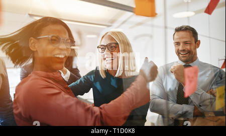 Two ecstatic young businesswomen laughing while brainstorming with sticky notes on a glass wall with colleagues in a modern office Stock Photo