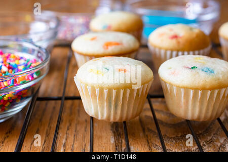 Mini cupcakes on wire rack waiting to be frosted.  Various colored sugars and sprinkles in glass bowls around cupcakes. Stock Photo