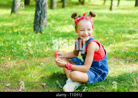 happy little girl in red T-shirt and headscarf in denim overalls holding ripe watermelon and laughing. the child is sitting on the grass in the park Stock Photo