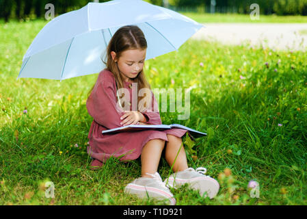 little girl in burgundy dress reading book in park under an umbrella on bright sunny day Stock Photo