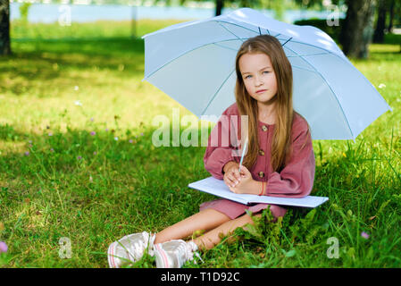 little girl in burgundy dress sits in park on grass in bright sunny day and reads book under umbrella Stock Photo