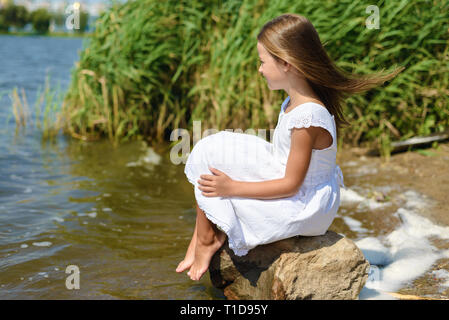little girl is sitting on rock in white dress on sunny day near river Stock Photo