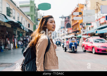 Young happy Asian female tourist backpacker walking and looking away on Khao San road in summer during trip to Bangkok, Thailand Stock Photo