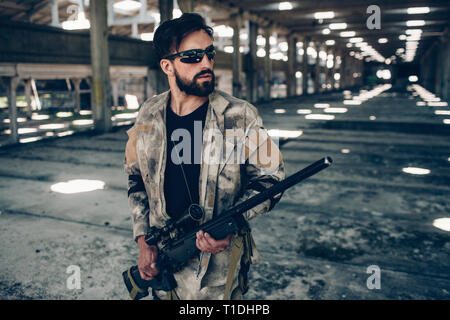 Attractive and serious brunette in uniform is standing and posing. He has paintball rifle in hands. Bearded man is looking straight forward. Stock Photo