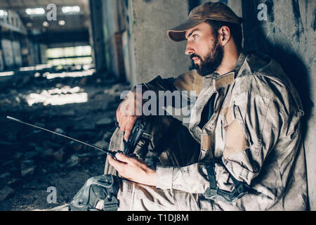 Thoughtful guy in uniform is sitting on the ground and leaning to wall. He is looking straight forward. Warrior is tired. Man is holding black guy and Stock Photo
