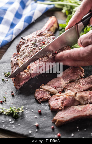 Freshly grilled tomahawk steak on slate plate with salt pepper rosemary and parsley herbs. Chef with knife cuts juicy bovine steak Stock Photo