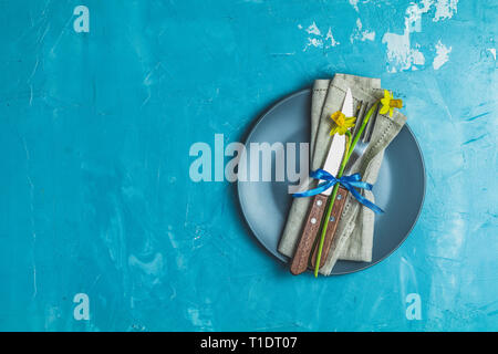 Empty blue plate and cutlery with daffodils on a napkin. Top view, green concrete surface background, copy space for you text Stock Photo