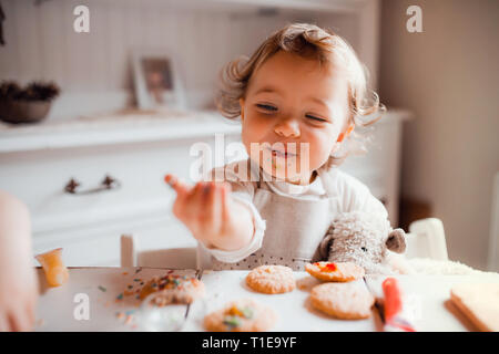 A small toddler girl sitting at the table, decorating cakes at home. Stock Photo