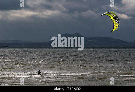 East Lothian, Scotland, United Kingdom, 26 March 2019. UK Weather: Kitesurfing at Longniddry Bents. A solitary kitesurfer with a bright yellow kite takes advantage of the stiff breeze in the Firth of Forth with the distinctive outline of Arthur's Seat in Edinburgh across the bay Stock Photo