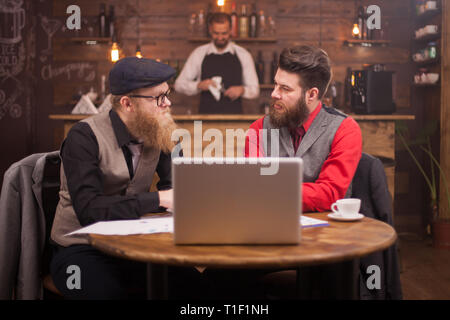 Two handsome businessmen looking at each other while talking about finances in a vintage pub. Stylish beard. Coffee cup. Laptop Stock Photo