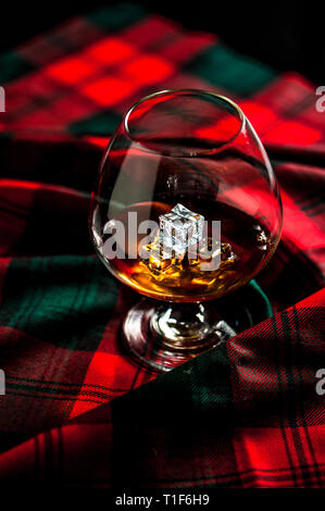 Scottish whiskey in a glass with ice on traditional red with green tartan. Crustal decanter and dry thistle. Selective focus. Stock Photo
